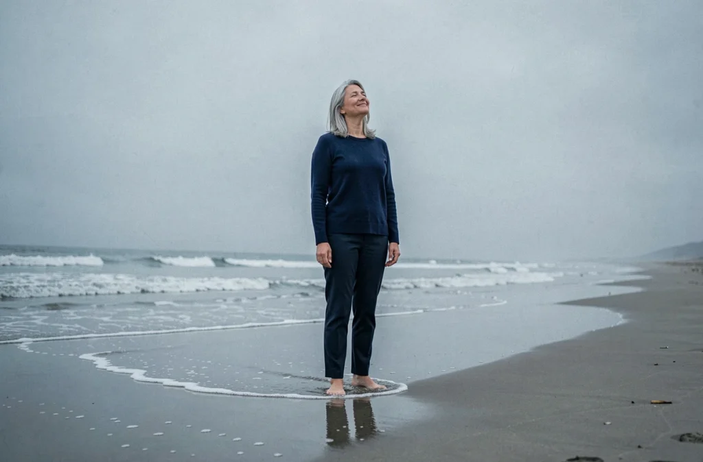 Femme sereine les pieds dans l'eau sur une plage brumeuse, symbolisant l'acceptation du moment présent et la vraie signification du carpe diem
